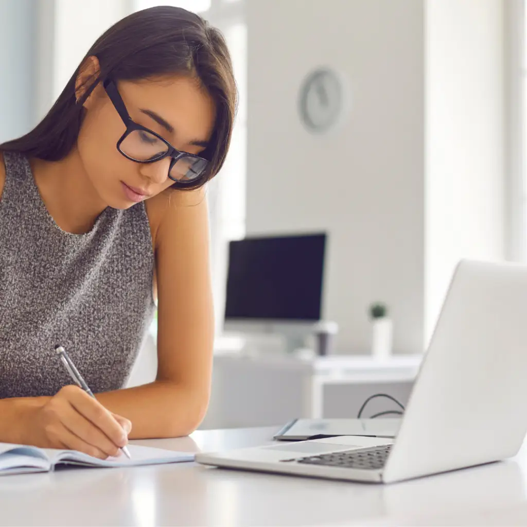 Woman with glasses writing in notebook while viewing laptop screen at desk
