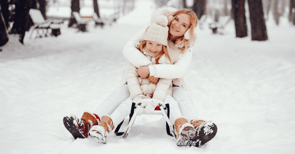 A mother and daughter embracing the outdoors in winter, sitting on a sled in the snow.