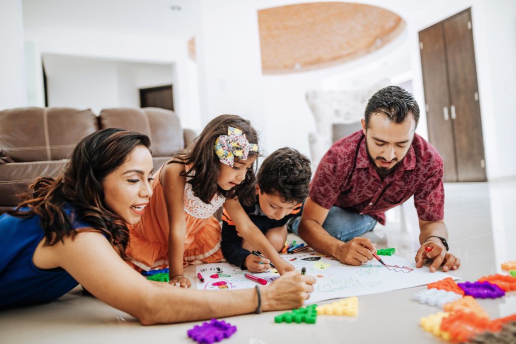 Happy family with two parents and two children lying on the floor, drawing and playing with colorful educational blocks together, promoting bonding, creativity, and child development.