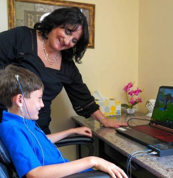 A woman is sitting in a chair with a boy in front of a laptop, conducting clinical research on mood and behavior.